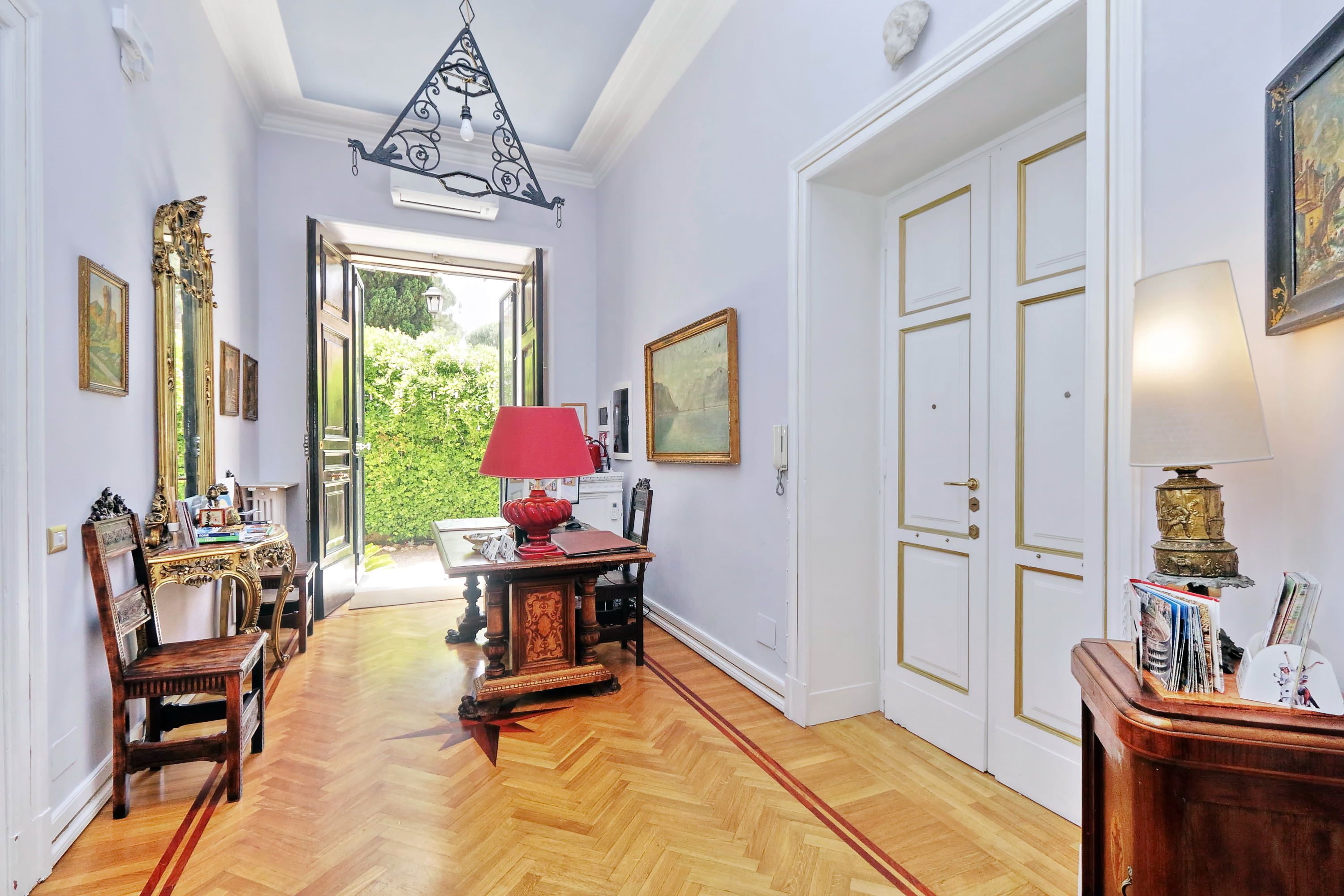 A hallway with wooden flooring, a red table lamp, and a desk with chairs. There are framed pictures on the walls and a door with a gold trim.