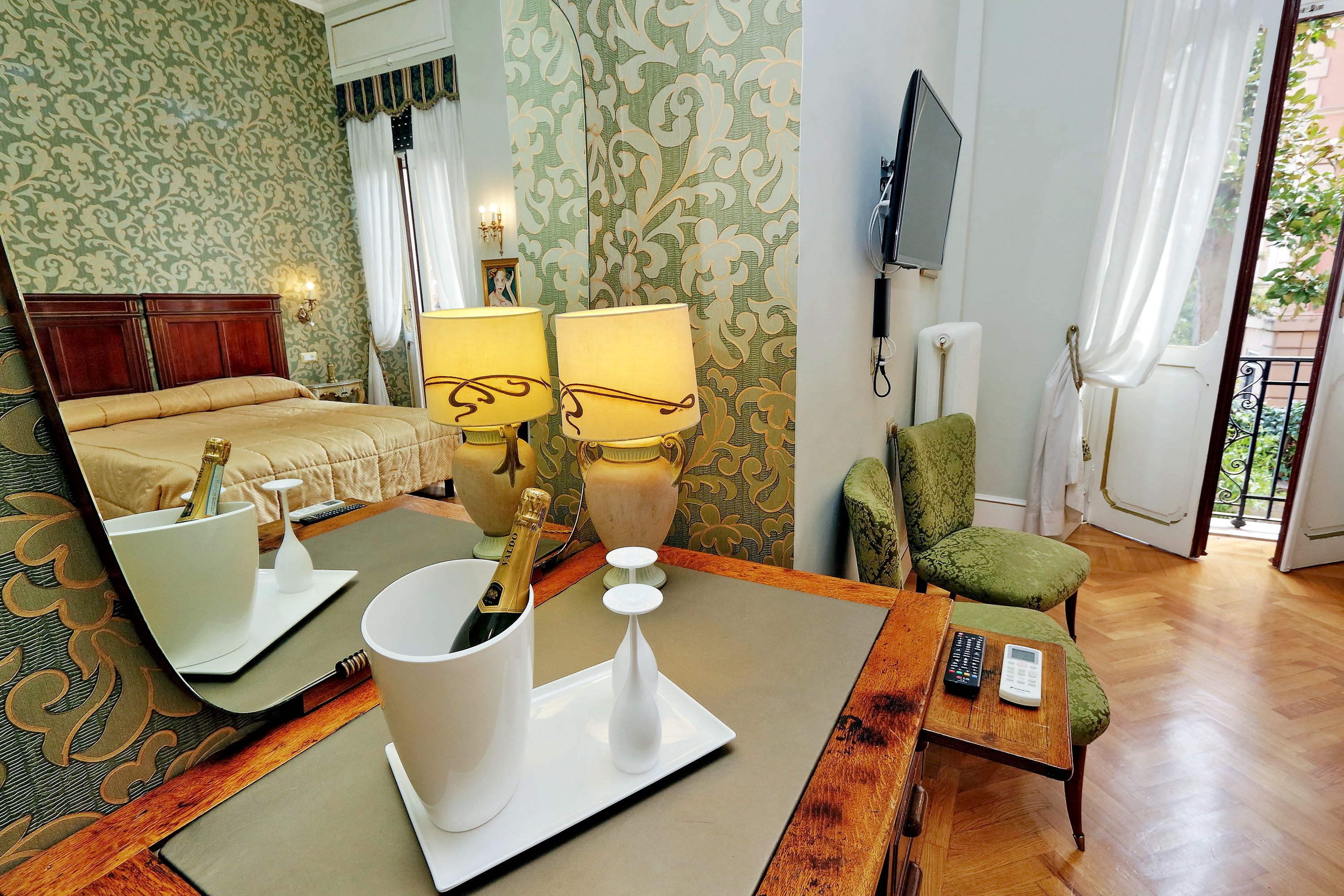 A hotel room with a bed, two bedside lamps, a green armchair, a small table with a remote control, and a mirror reflecting the room.