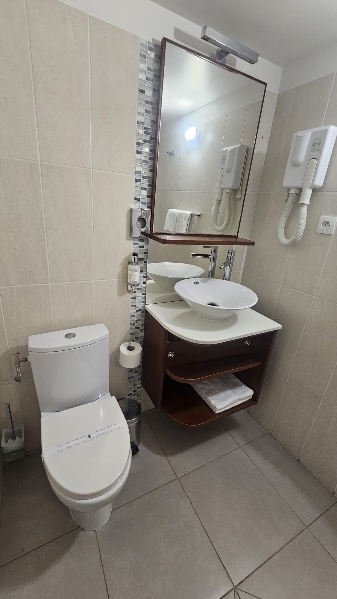 Un baño con inodoro, dos lavamanos, un espejo, toallero y secador de manos.