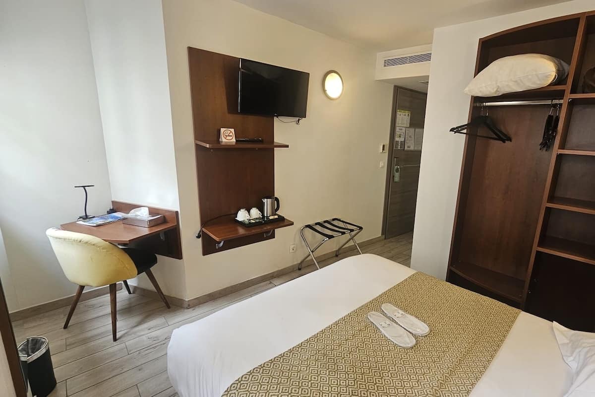 A hotel room with a bed, a desk with a chair, a TV mounted on the wall, a wardrobe, and a small table with a kettle and cups.