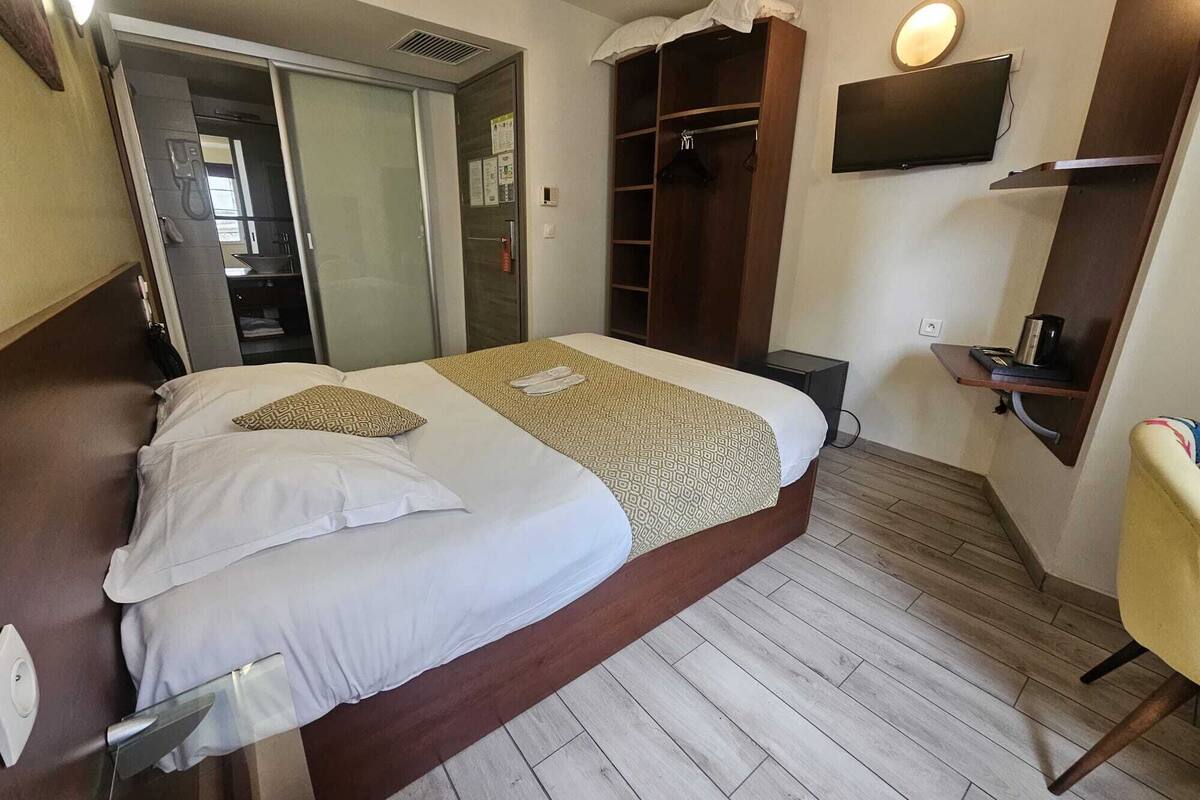 A hotel room with a bed, a wardrobe, a TV mounted on the wall, and a small table with a chair.