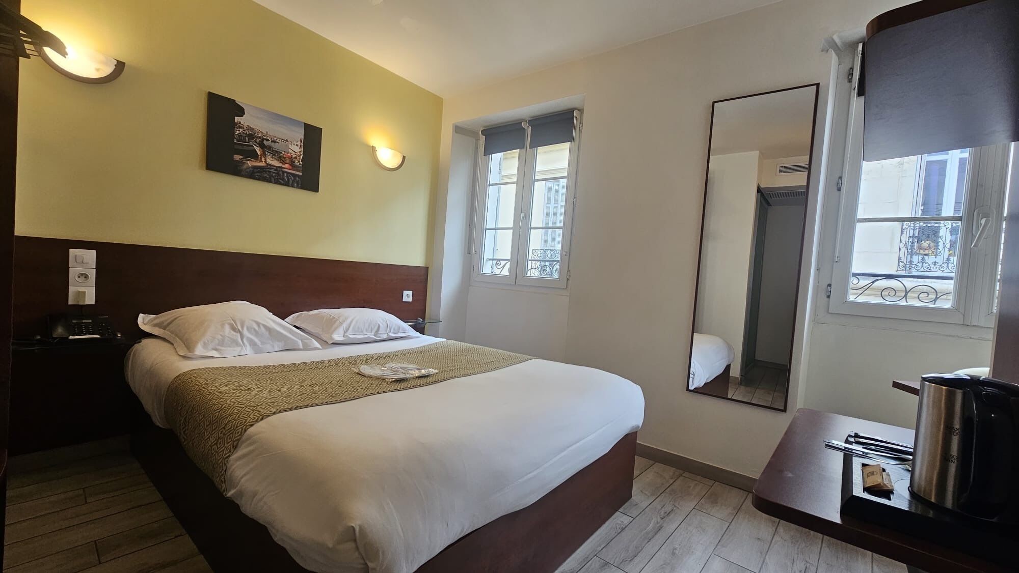 A hotel room with a bed, bedside table, lamp, window, and a mirror.