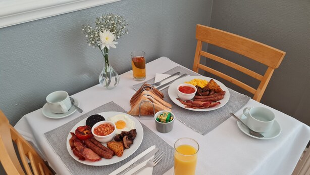A breakfast table with plates of eggs, bacon, sausages, beans, and toast, a glass of orange juice, and a vase with flowers. - The Chimes (Bridlington)
