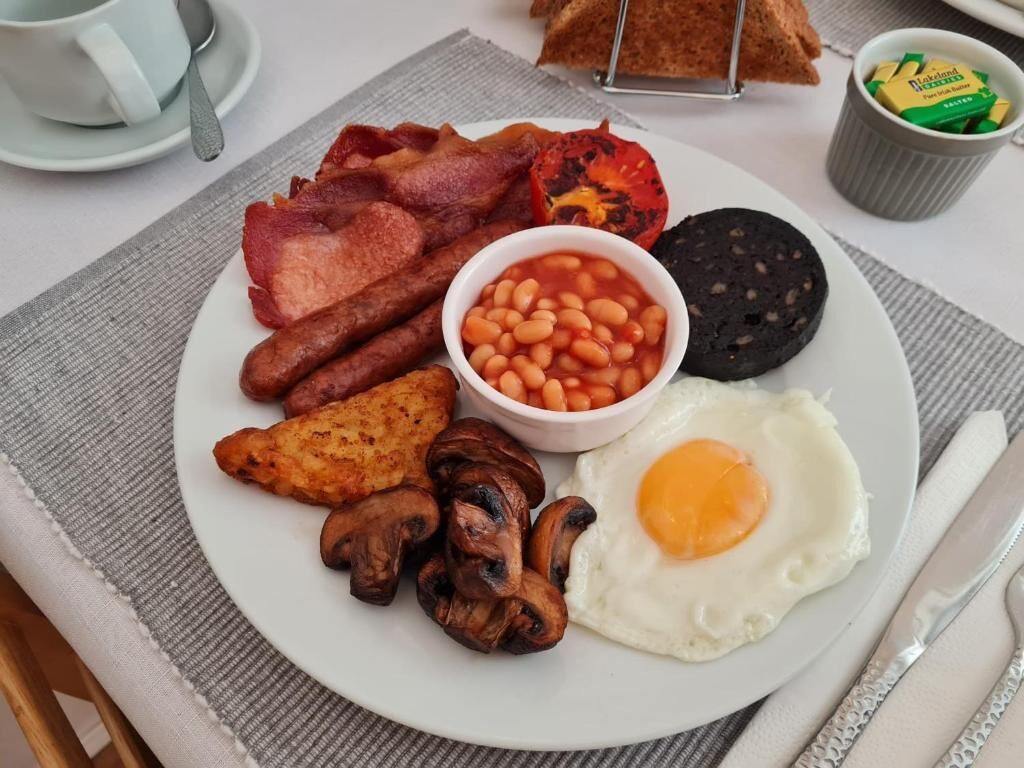 Un plato de comida para el desayuno con tocino, huevos, frijoles, champiñones y una rebanada de morcilla.