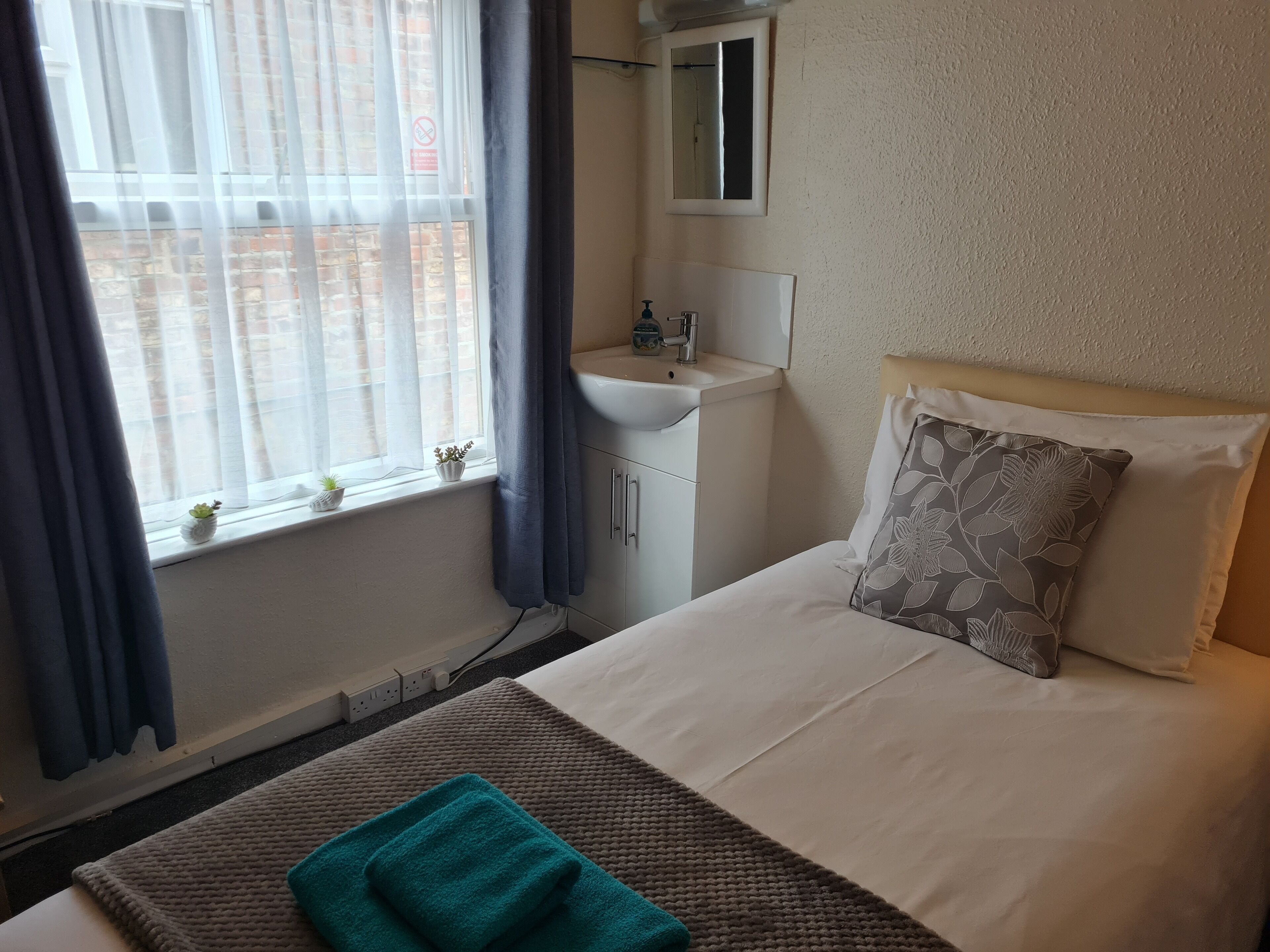 A hotel room with a bed, a bedside table, a window with curtains, a sink, and a mirror.