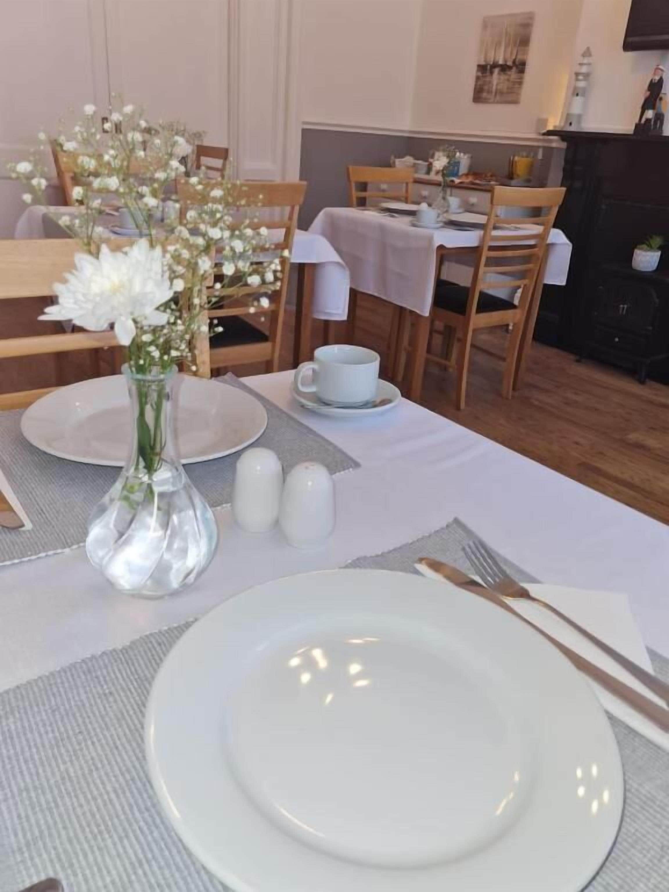 Une table à manger dressée avec de l’assiette blanche, un vase en verre transparent contenant des fleurs, des salières et des poivrières, et une tasse.