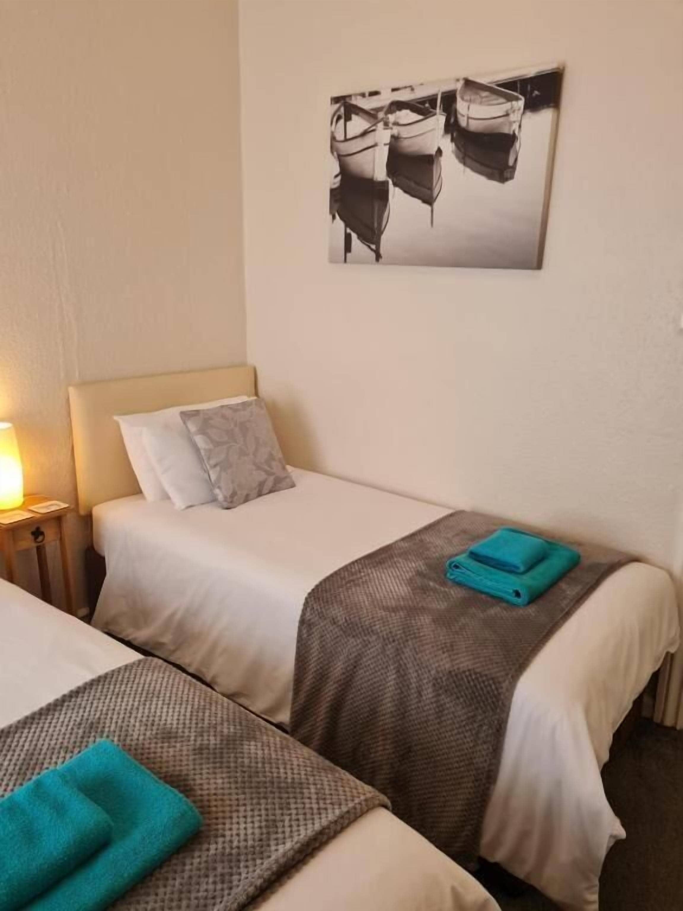 A hotel room with two beds, each with a pillow and a blanket. There is a wall-mounted picture above the beds.