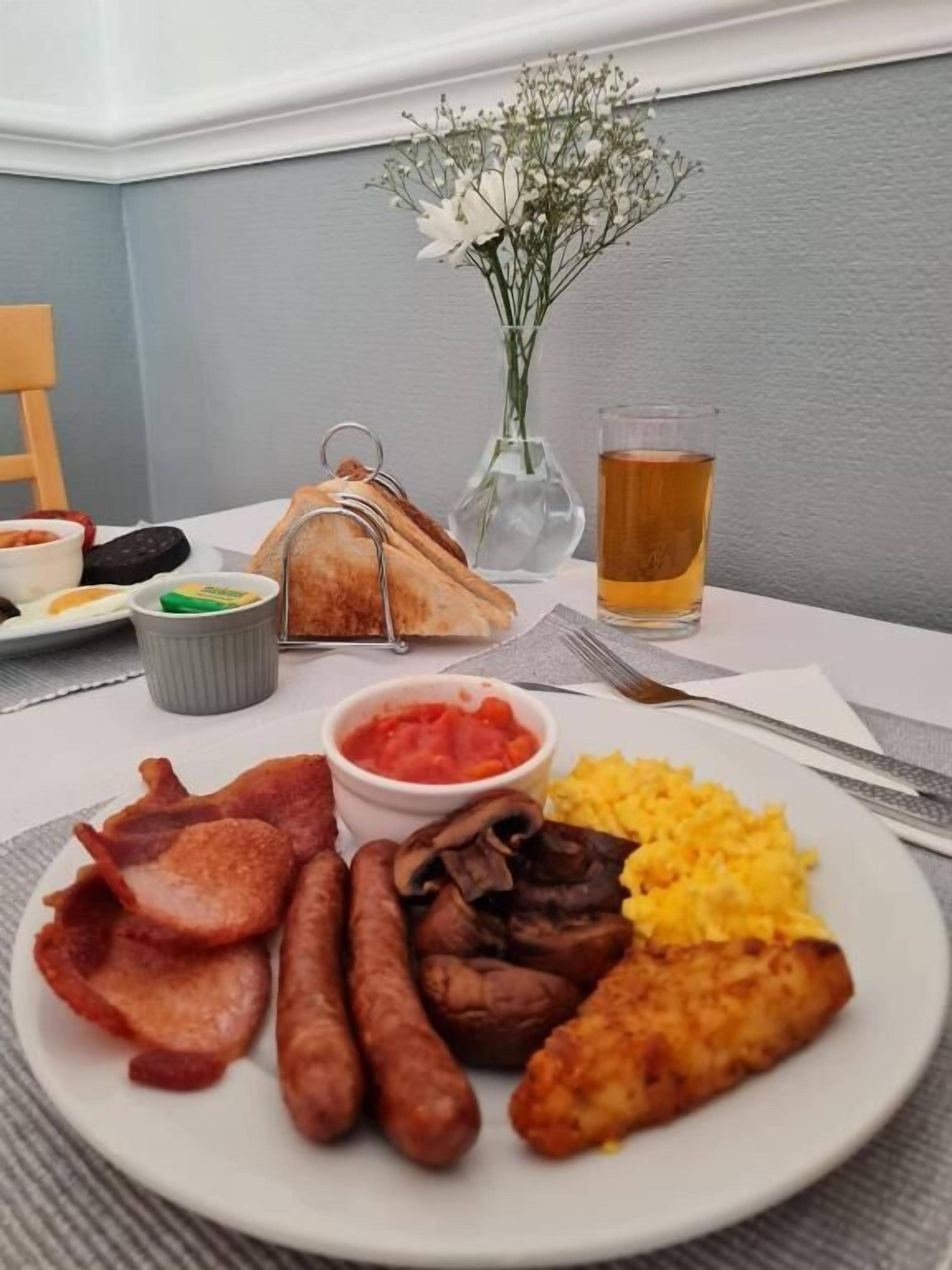 A breakfast plate with sausages, fried fish, and scrambled eggs, accompanied by a glass of tea and a vase of flowers.
