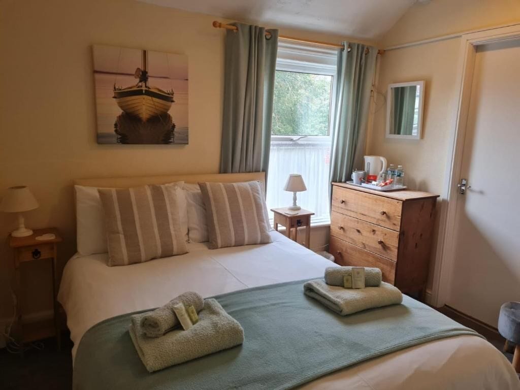 A hotel room with a bed, a dresser, a window with curtains, a painting on the wall, and a door.