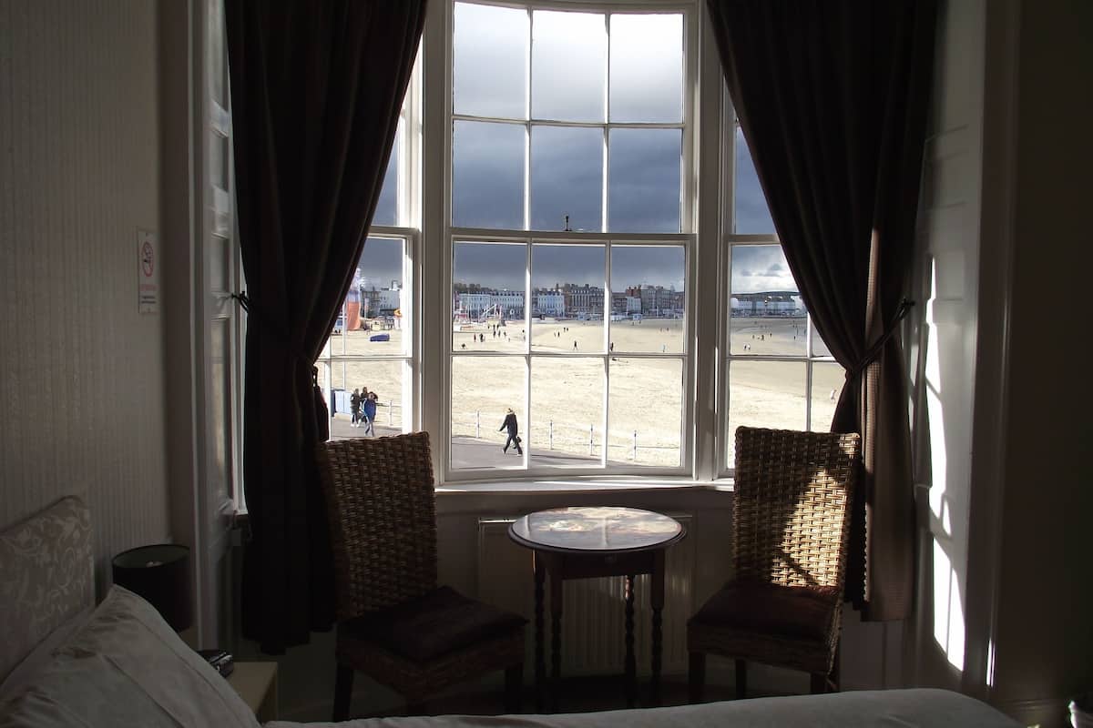 A room with a large window overlooking a beach, two wicker chairs, a small round table, and a bed with white linens.