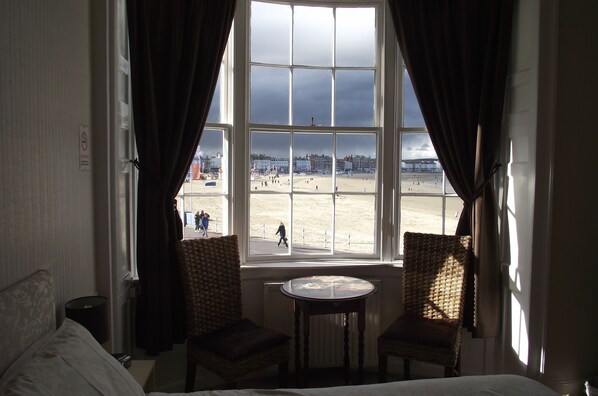 A room with a large window overlooking a beach, two wicker chairs, a small round table, and a bed with white linens. - The Anchorage (Weymouth)