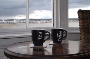 Two black mugs with "tea" and "coffee" on them, placed on a round wooden table by a window overlooking a beach. - The Anchorage (Weymouth)