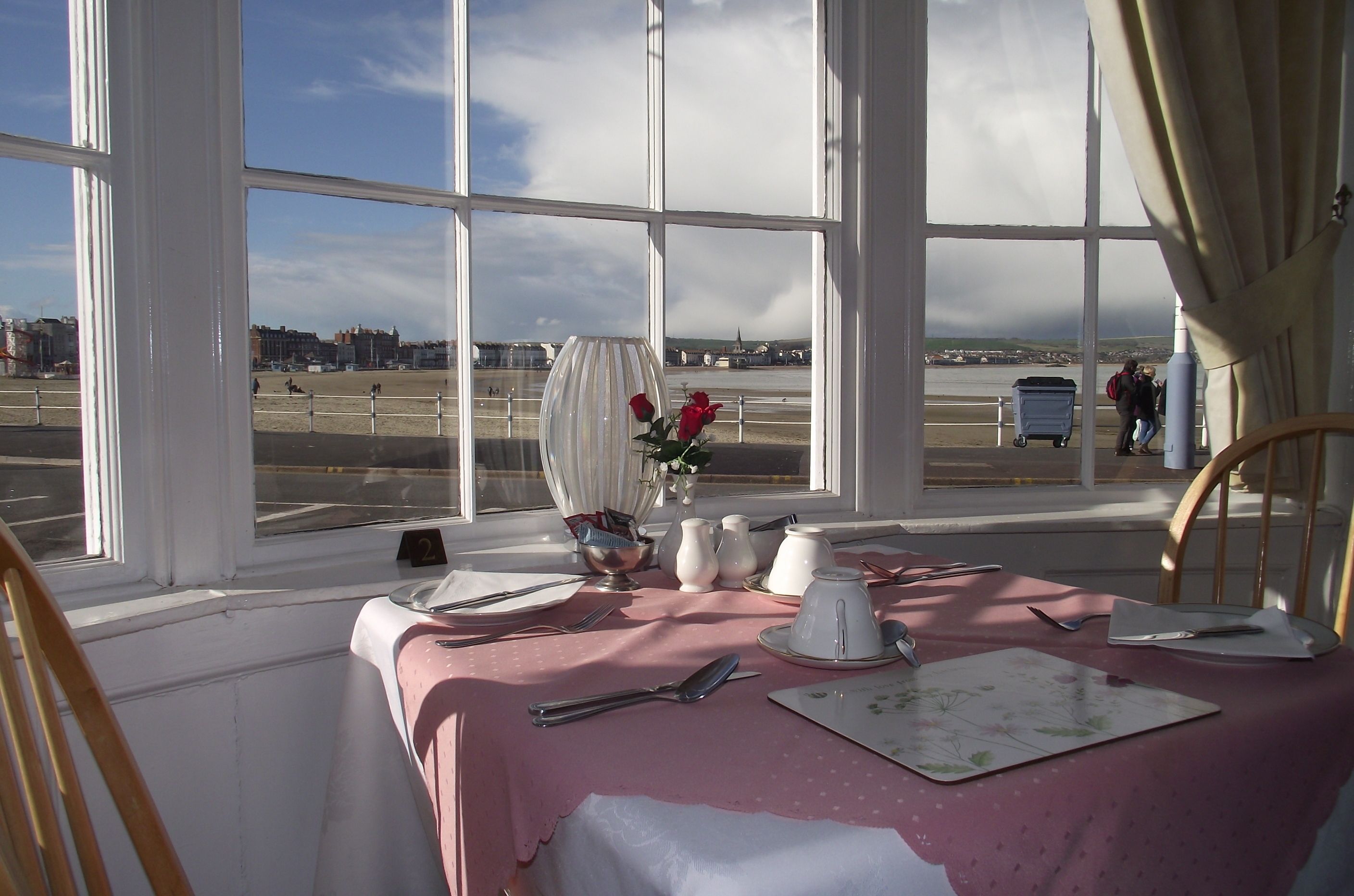 A dining table set with a pink tablecloth, white plates, silverware, and a vase with red flowers. Large windows offer a view of a beach and a distant cityscape.