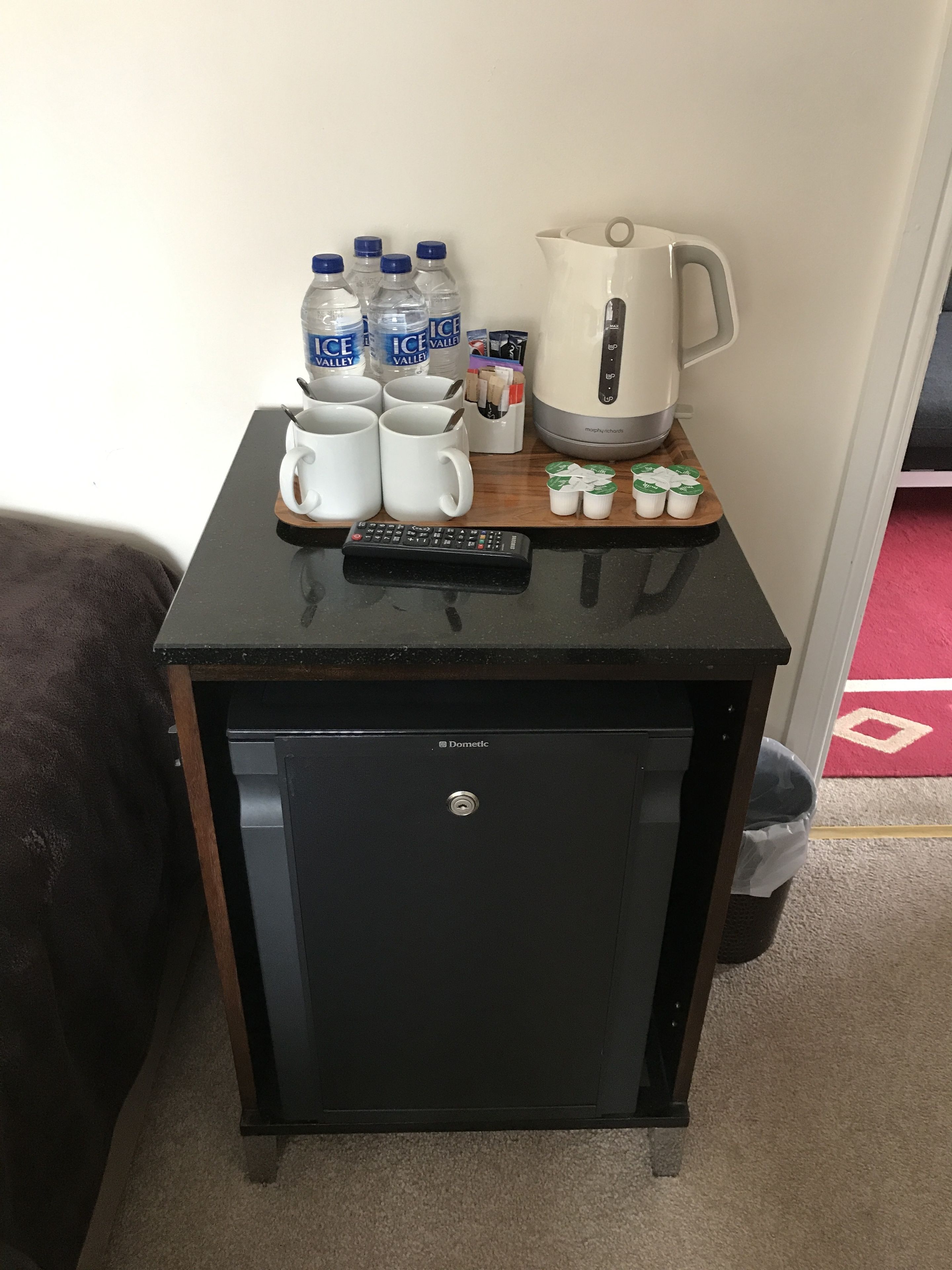 Een klein zwart kastje met een afstandsbediening erop, waarin een waterkoker, waterflessen en een mokset zitten.