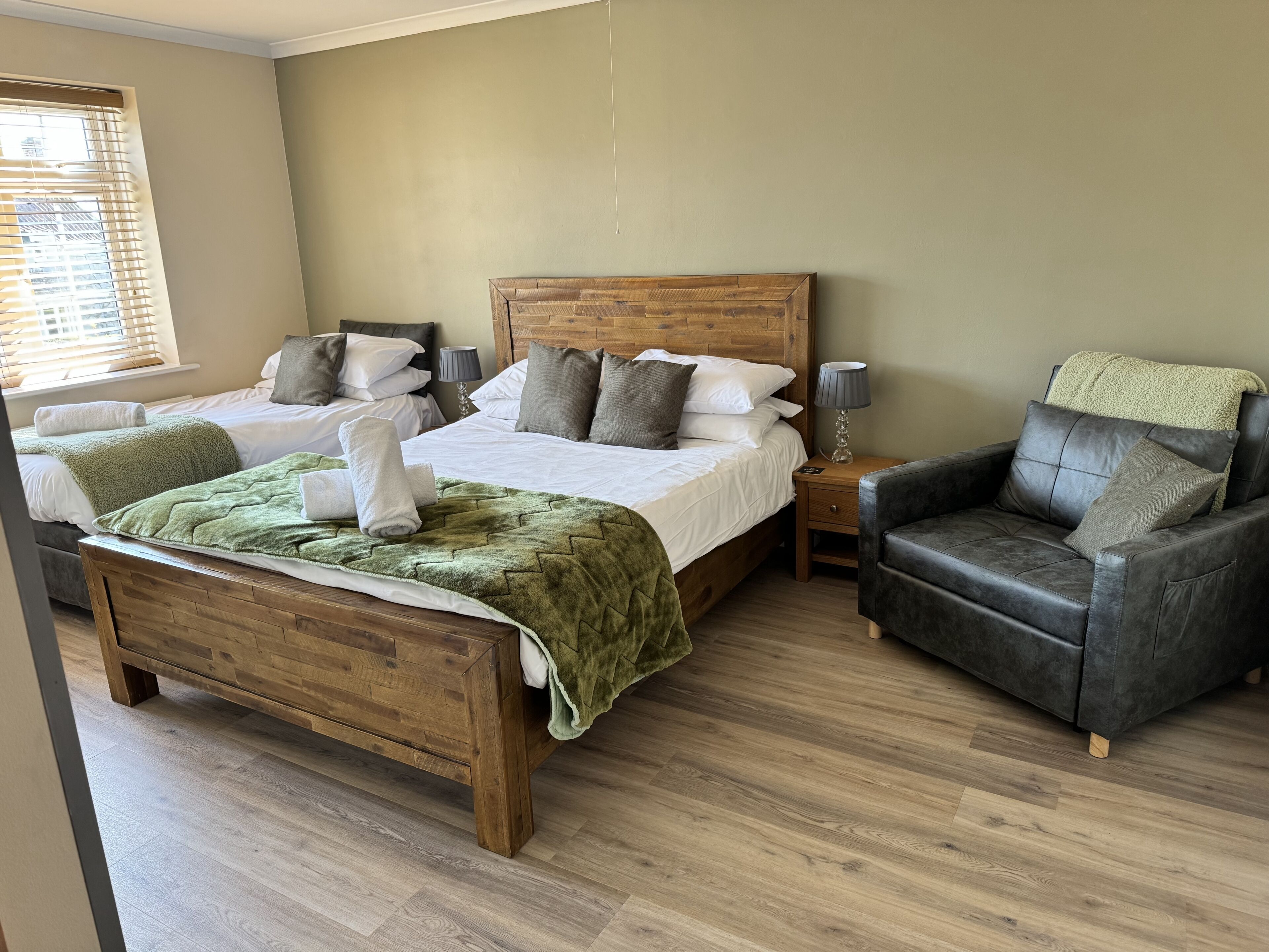 A hotel room with a large bed, a smaller bed, a wooden headboard, a leather armchair, and a bedside table with a lamp.