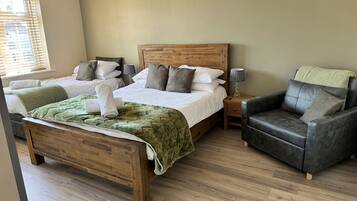 A hotel room with a large bed, a smaller bed, a wooden headboard, a leather armchair, and a bedside table with a lamp.