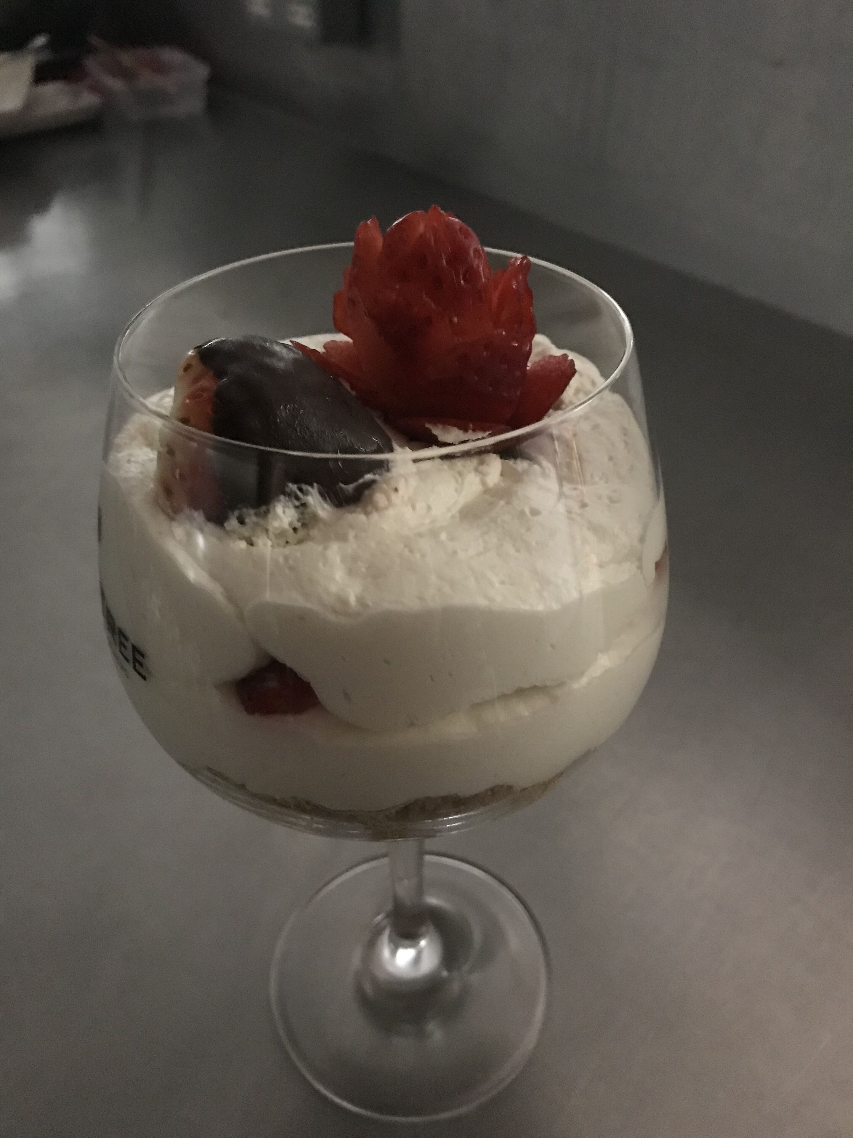 A dessert in a clear glass with a spoon, containing whipped cream, strawberries, and a dark fruit.