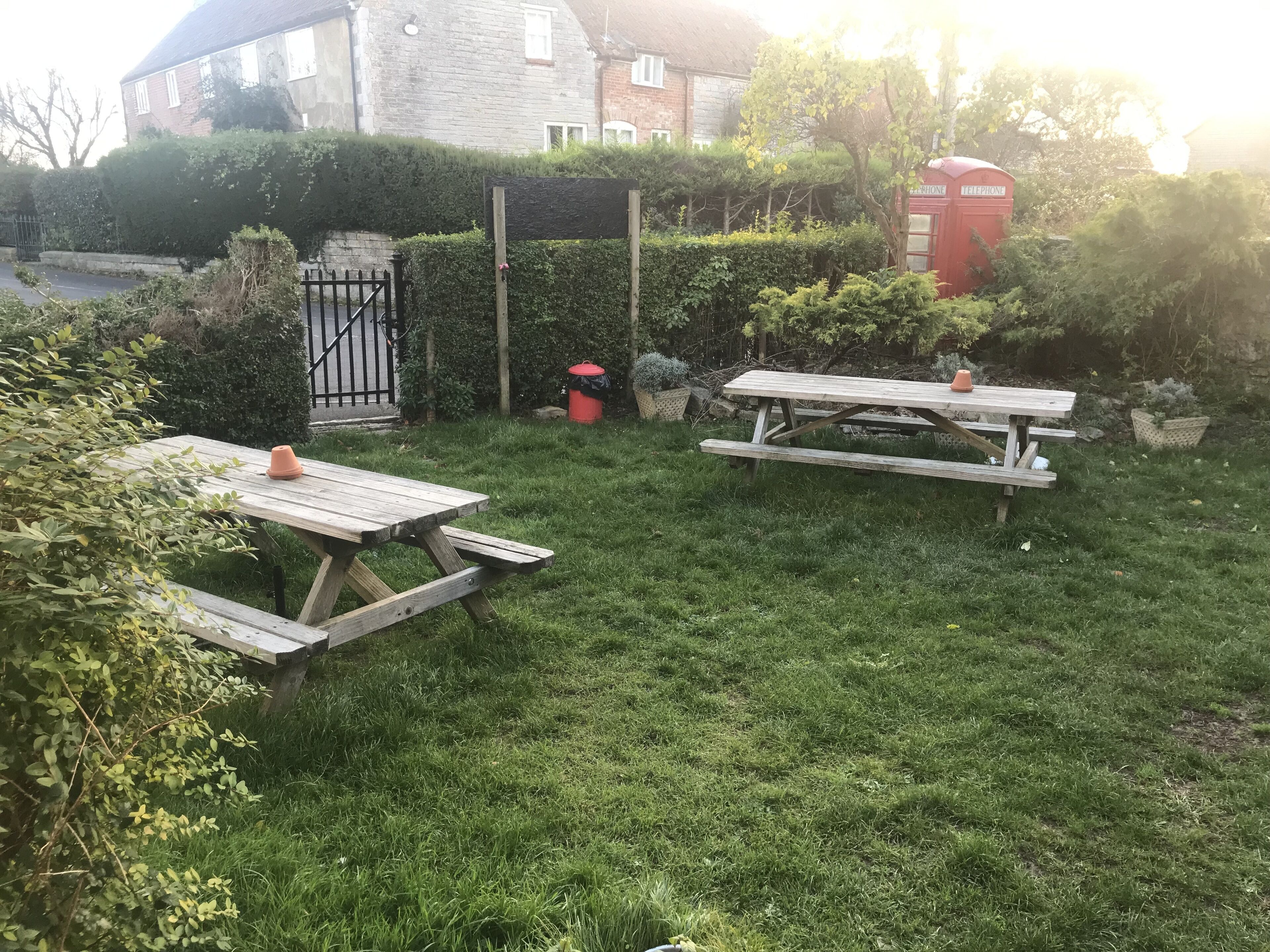 Un patio trasero con césped, dos mesas de picnic de madera, una cabina telefónica roja y un edificio de piedra al fondo.