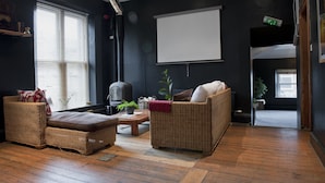 A cozy living room with wicker furniture, a fireplace, and a large window with blinds. - CellB - Hostel (Blaenau Ffestiniog)