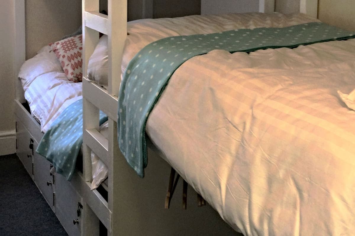 Uma cama box com beliches, um embaixo e um em cima, ambos com roupa de cama branca. Há gavetas para guardar objetos embaixo do beliche inferior.