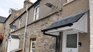A stone building with a white door marked '3', a wooden bench, and a yellow waste bin. - 3 Varis Apartments (Forres)