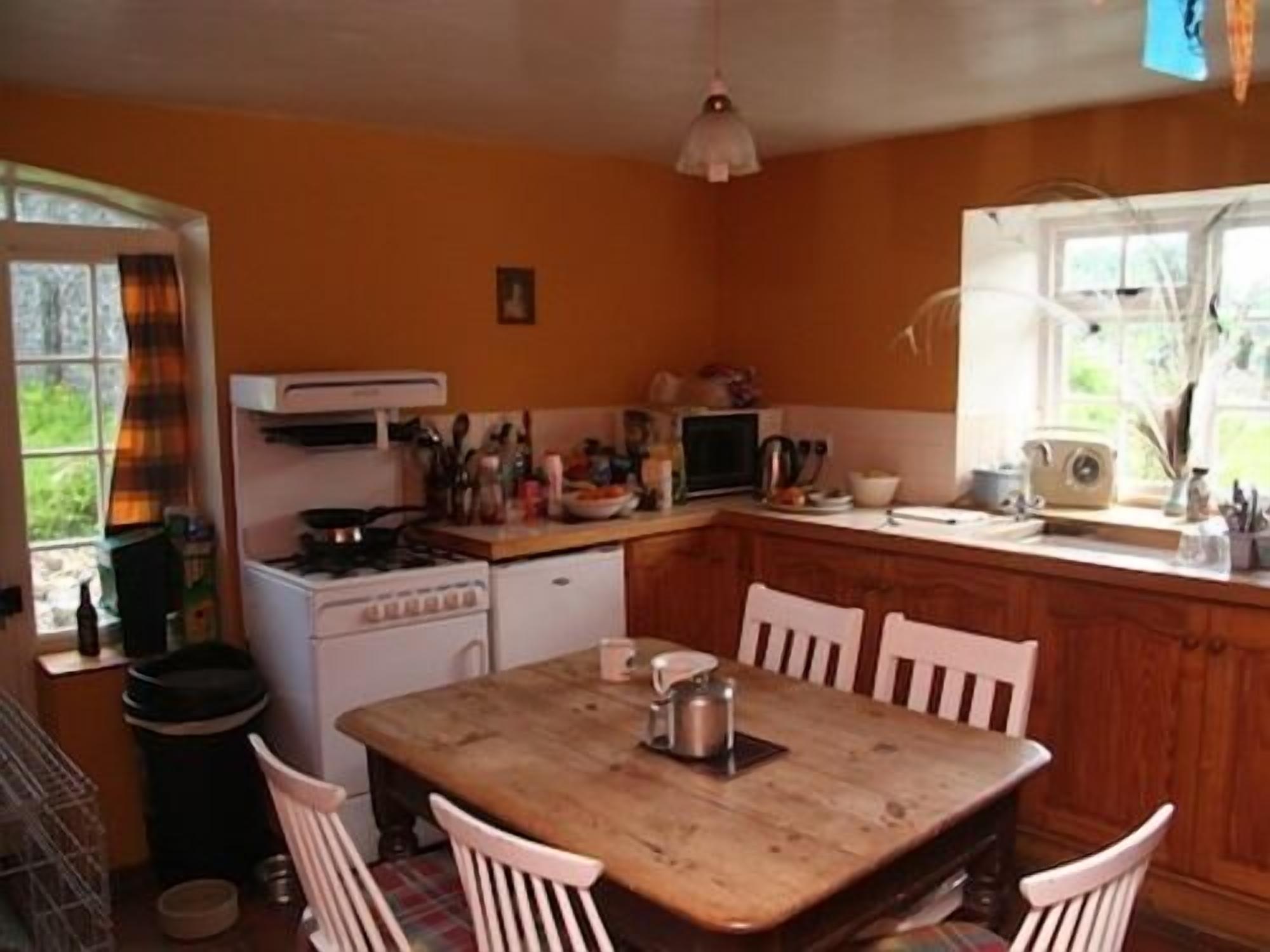 A compact kitchen with a wooden table and white chairs, a stove, a microwave, and a window with a view of greenery.