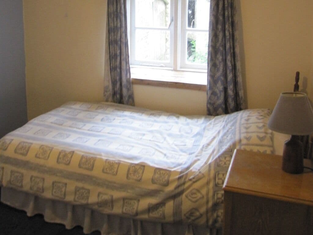 A small bedroom with a bed, a nightstand, a lamp, and a window with curtains.
