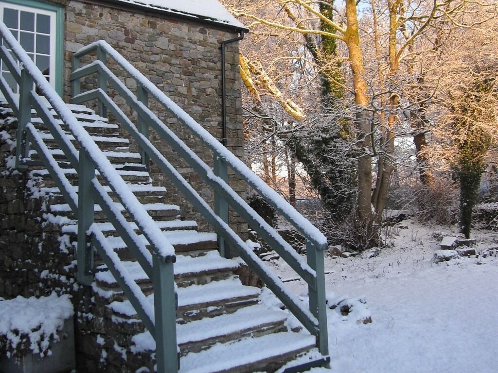 Un bâtiment en pierre avec un escalier menant à une porte, entouré d’un sol couvert de neige et d’arbres sans feuilles.