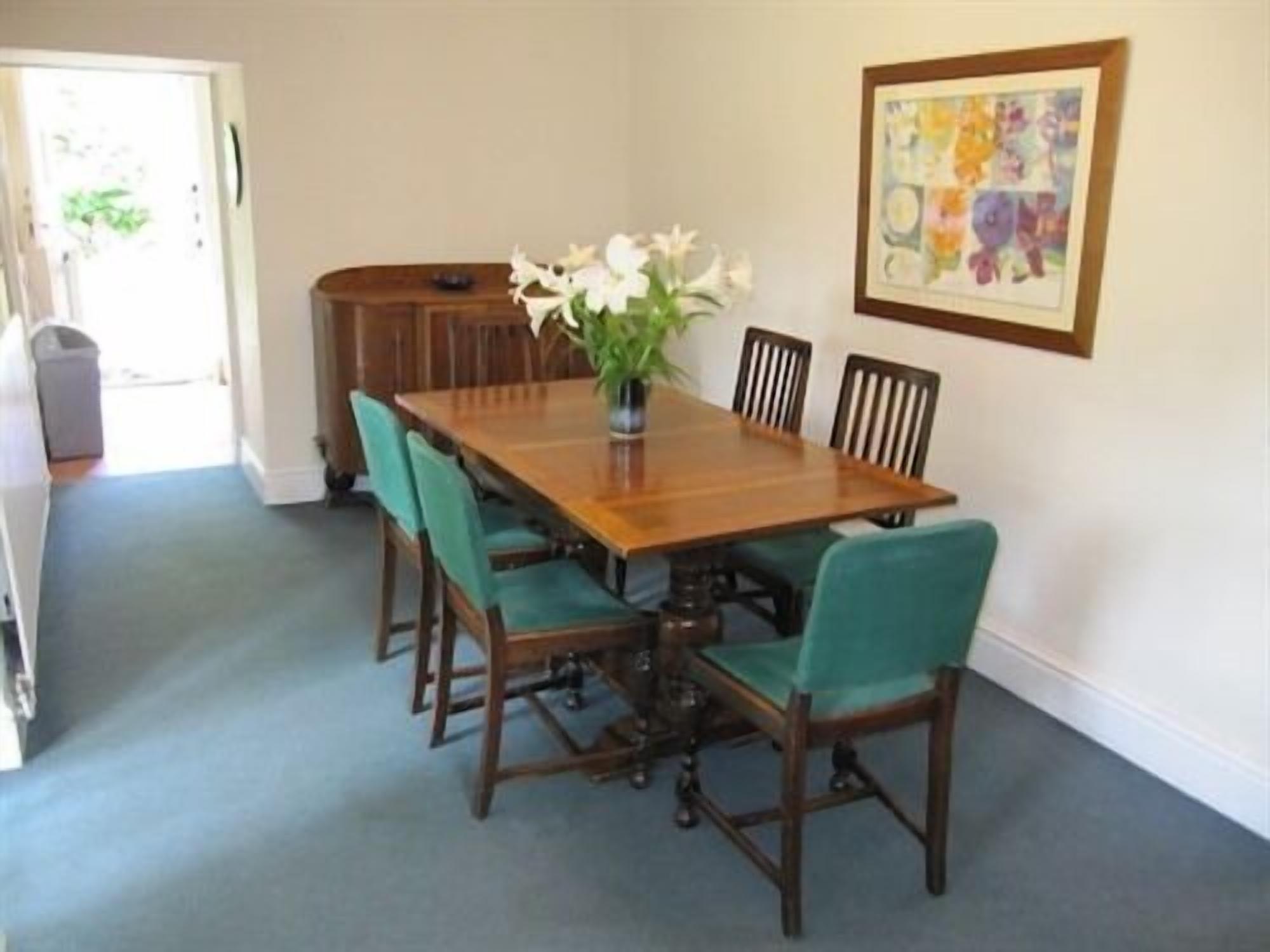 A dining area with a wooden table and six chairs, a vase with flowers, and a framed picture on the wall.