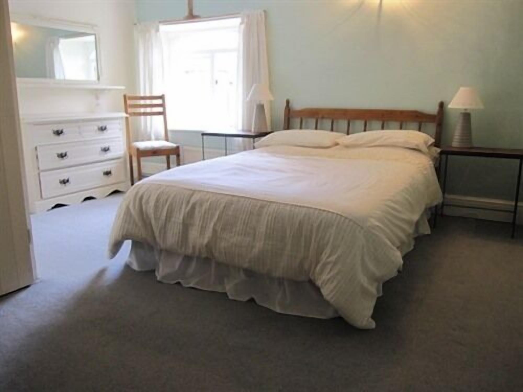 Een slaapkamer met een bed, een dressoir, een stoel, een nachtkastje en een lamp.