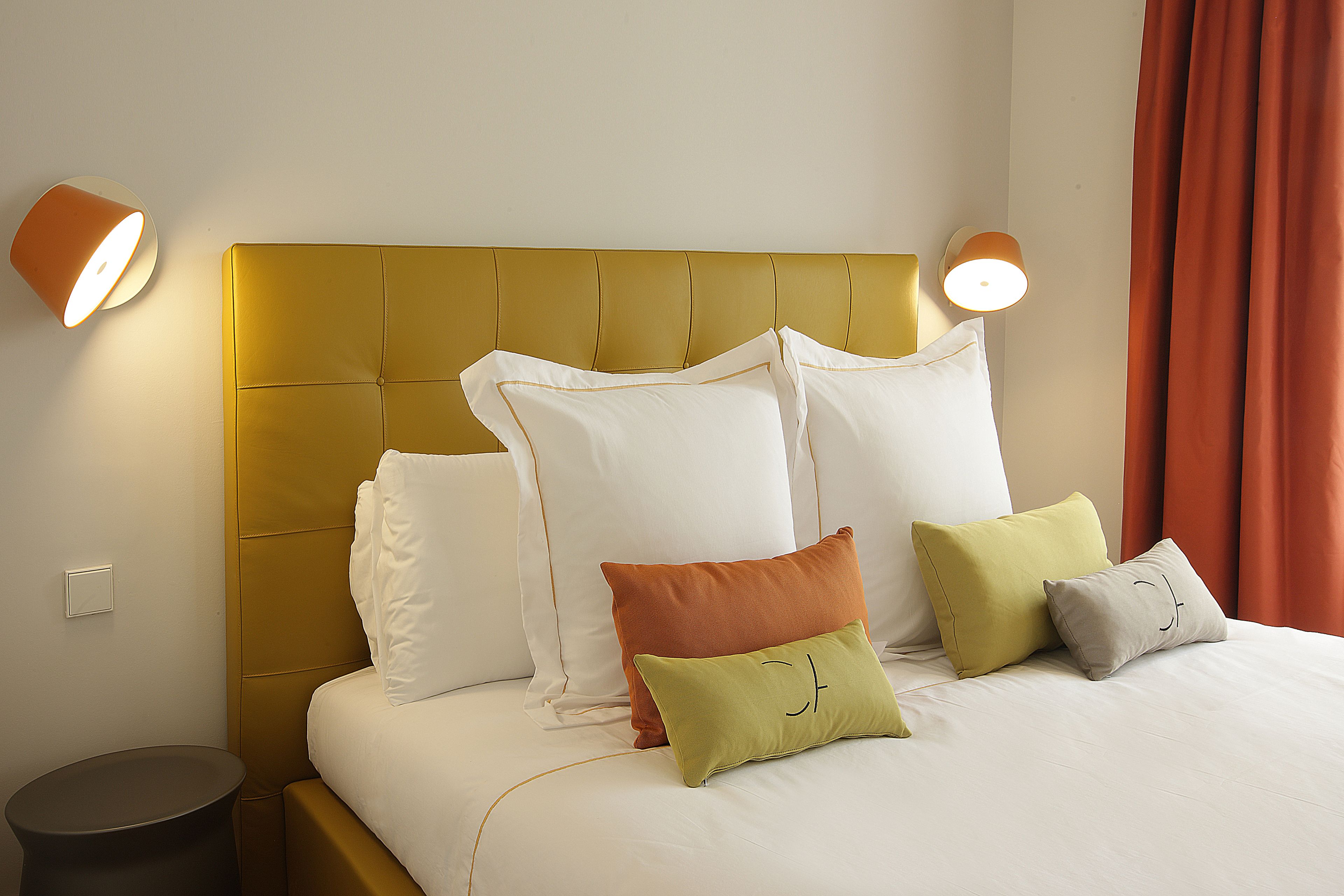A neatly made bed with a gold headboard, white and colored pillows, and two wall-mounted lamps.