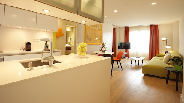 A modern kitchen with a white countertop, a sink, and a vase of yellow flowers. There is a dining area with a table and chairs, and a living room with a sofa and a TV. - Cosmo Apartments Rambla de Catalunya (Barcelona)