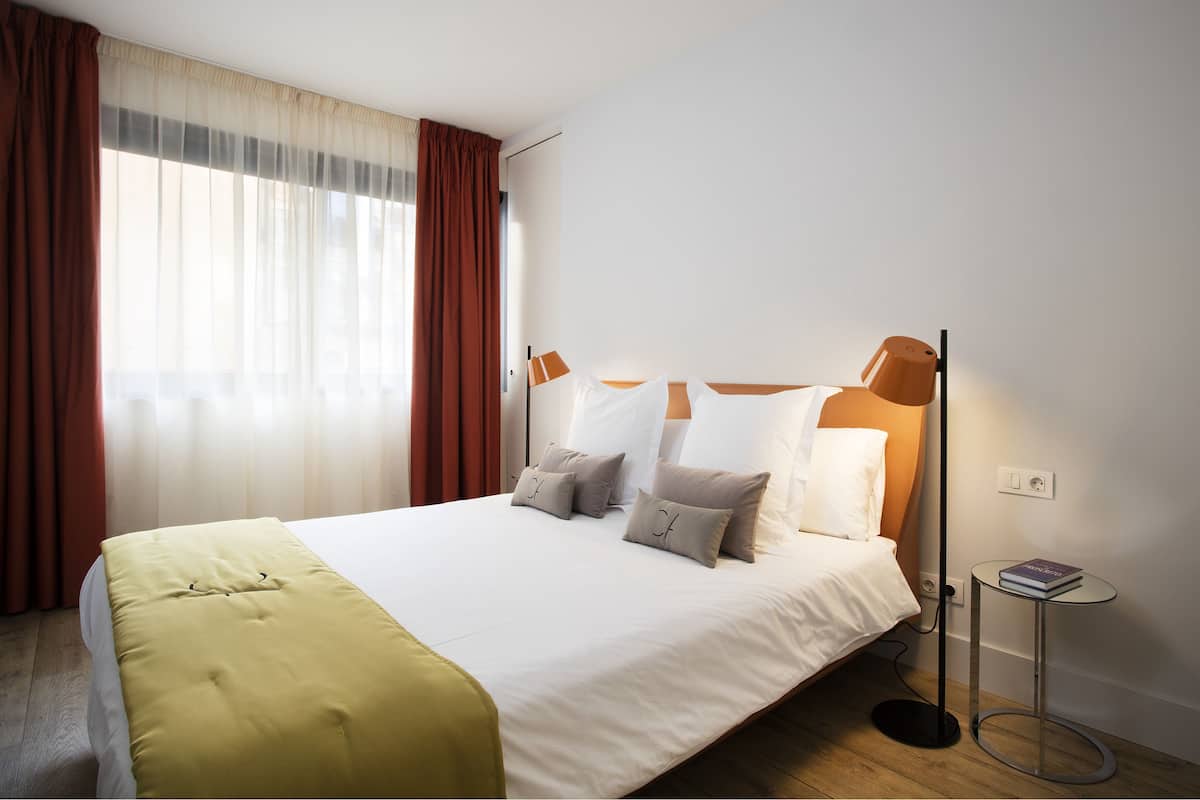 Un lit bien fait, recouvert d’une couvre-lit blanche et jaune, une table de chevet avec un livre, et une lampe sur pied.