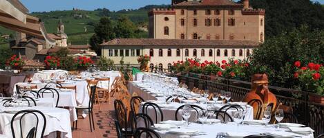 Un área de comedor al aire libre con mesas dispuestas para una comida, un edificio grande al fondo y vistas del paisaje circundante.