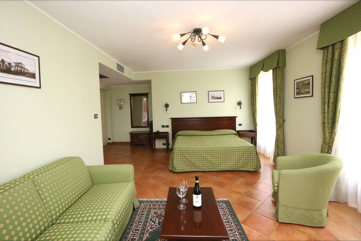 A hotel room with a green sofa, a bed with green bedding, a wooden table with a bottle and glass, and a ceiling fan.