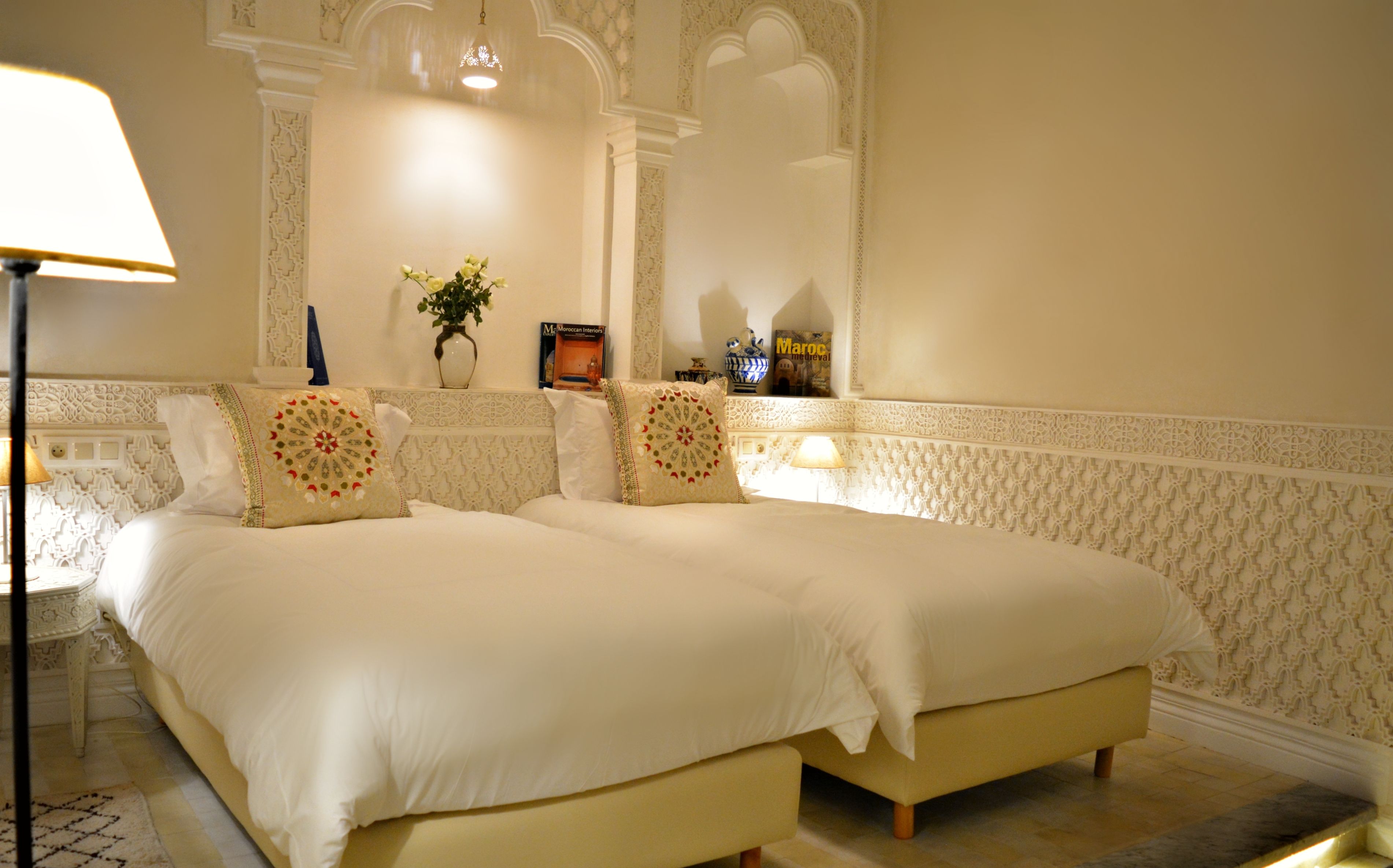 A hotel room with two beds, a nightstand, a lamp, a vase with flowers, and a book titled "Maroc medieval".