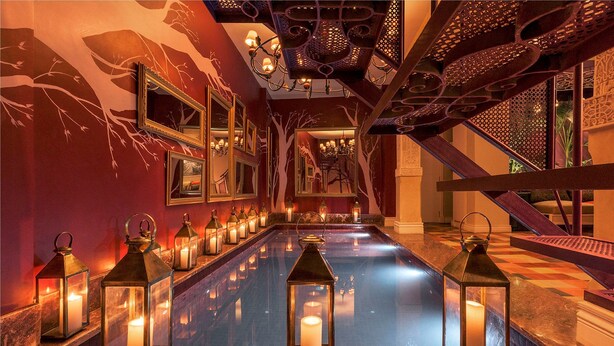 Un área de piscina interior lujosa con superficie reflectante, rodeada de ornamentales decoraciones en las paredes y varios espejos.
