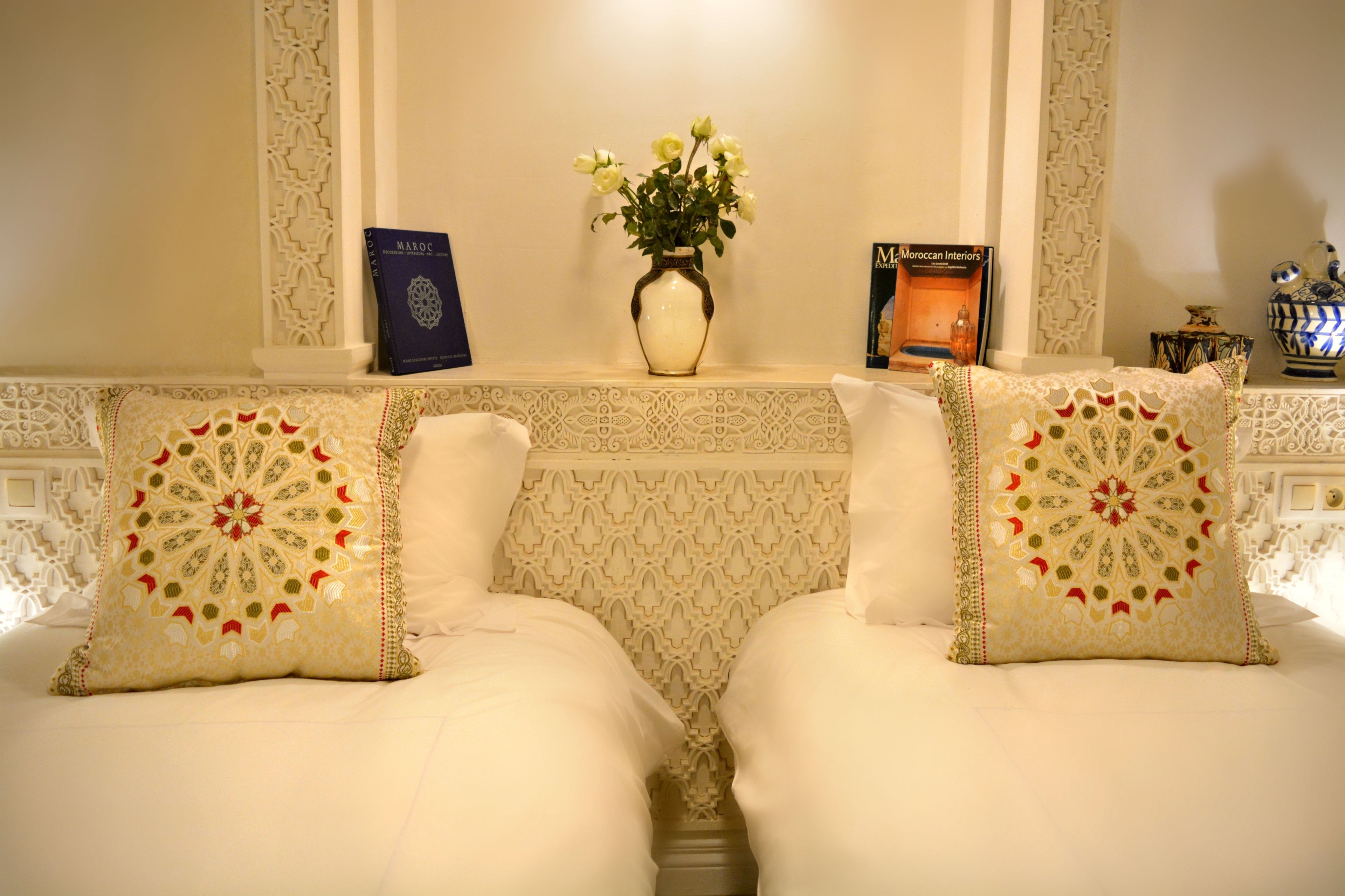 Een kamer met twee bedden, een vaas met bloemen, een boek getiteld 'MOROCCAN INTERIORS' en decoratieve kussens.