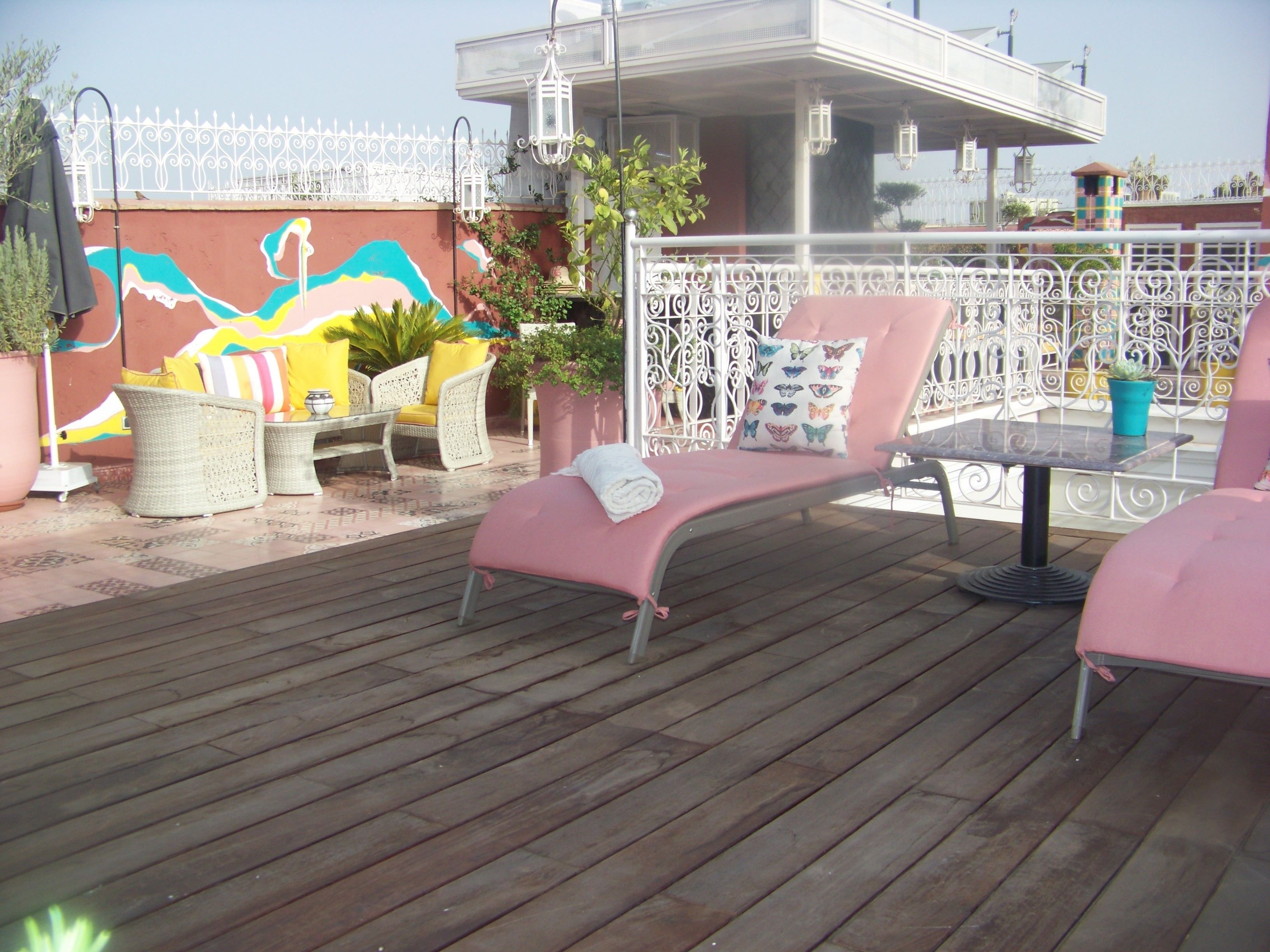 A wooden deck with outdoor seating, including a pink lounge chair and wicker chairs, surrounded by potted plants and a decorative fence.