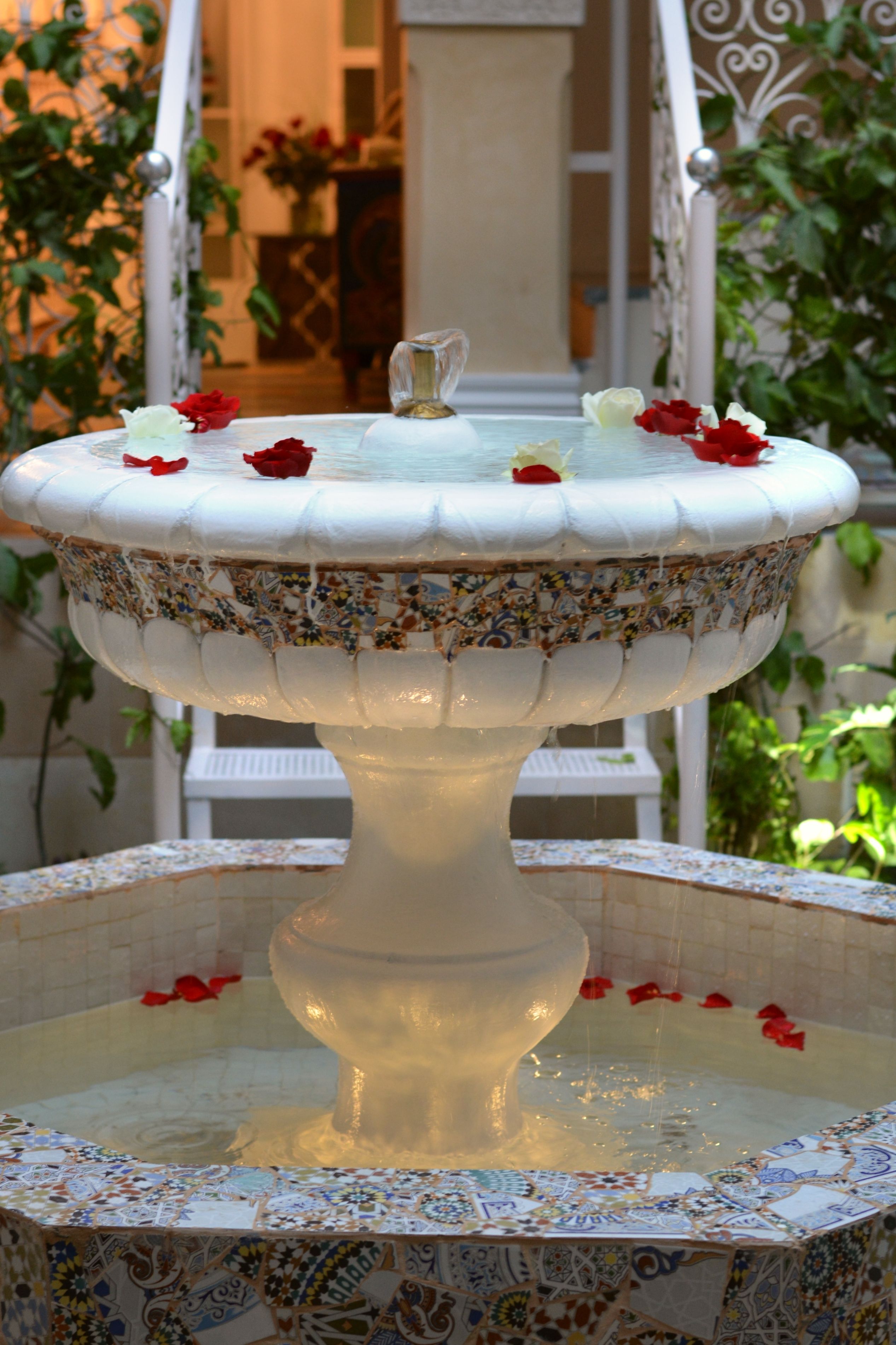 Une fontaine dotée d’une base en mosaïque et d’un piédestal blanc, entourée de verdure et agrémentée d’une chaise blanche.