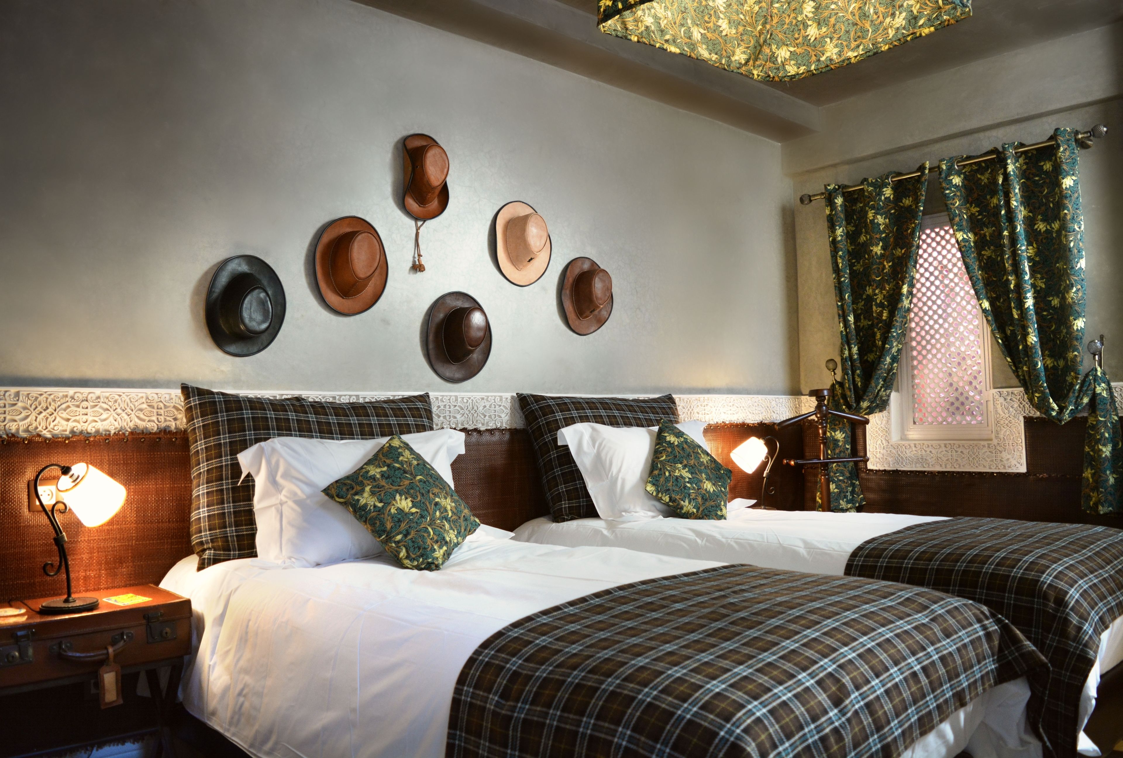 A hotel room with two beds, a wall-mounted hat display, a nightstand with a lamp, and a window with curtains.