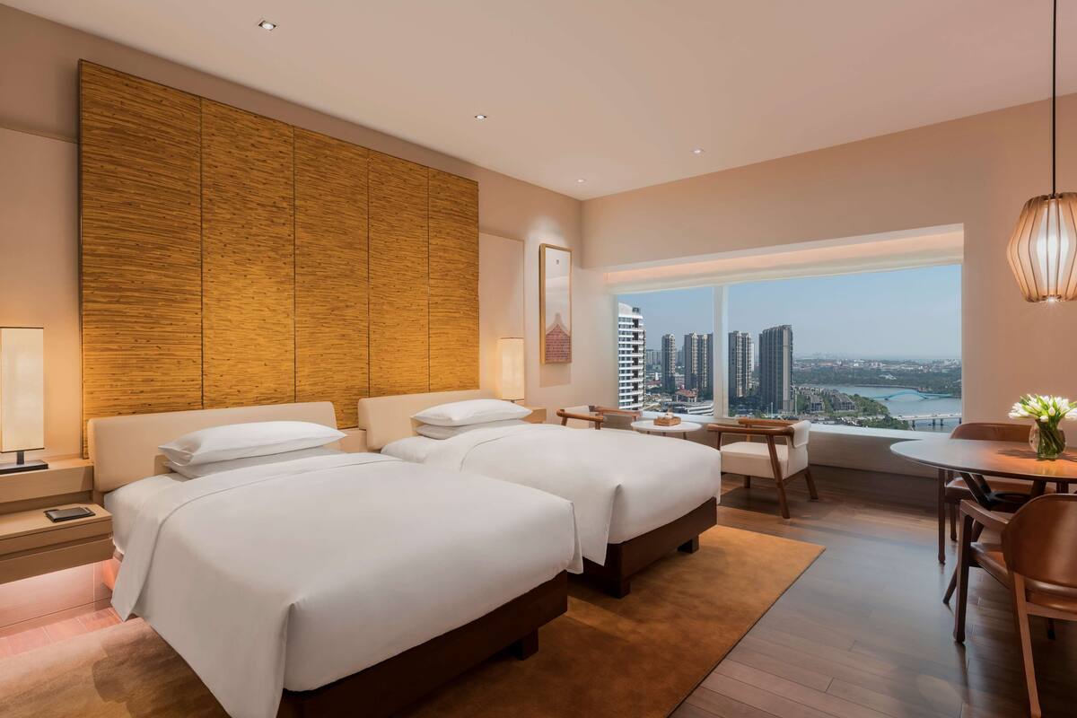 A hotel room with two beds, a dining area, and a large window with a city view.