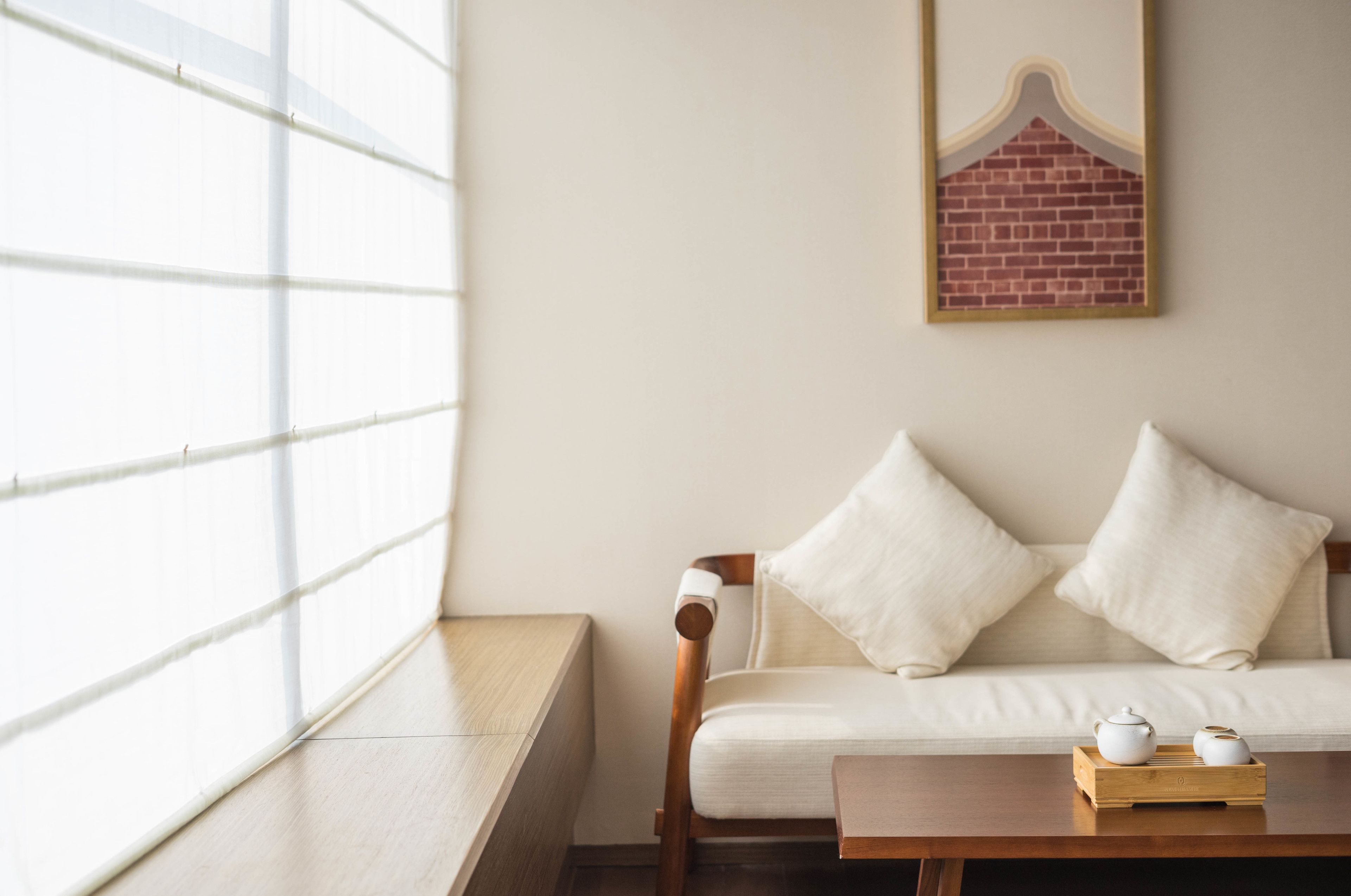 A room with a wooden bench, a white sofa with two pillows, a wooden table with two white teapots, and a window with blinds.