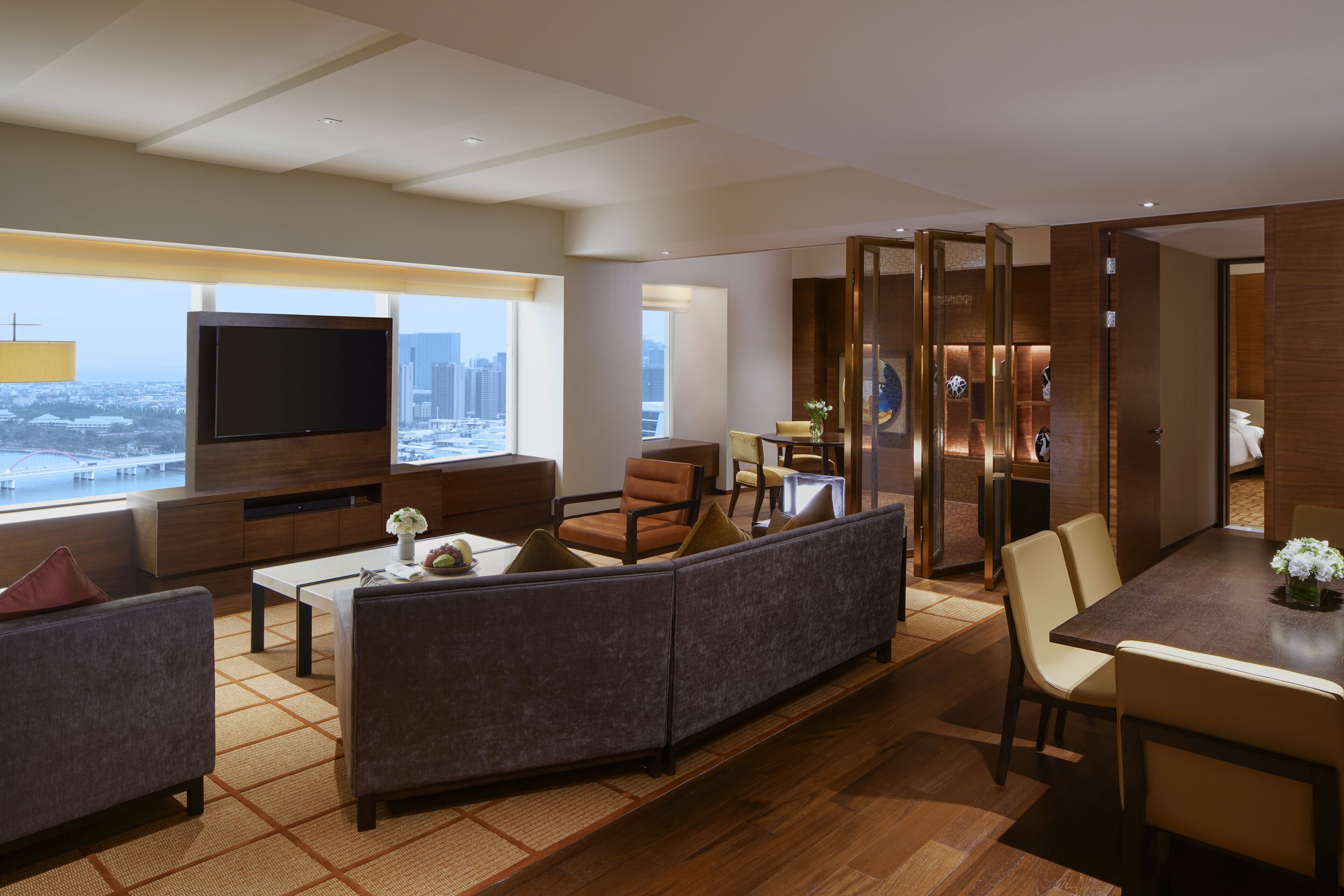 A modern hotel living room with a large TV, comfortable seating, a dining area, and a view of the city.