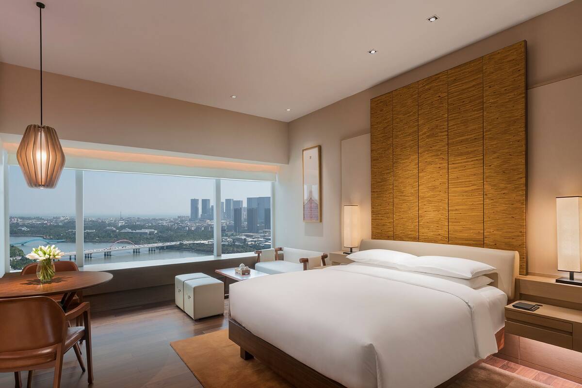 A modern hotel room with a large bed, a dining area with a table and chairs, a view of the cityscape, and a wooden accent wall.