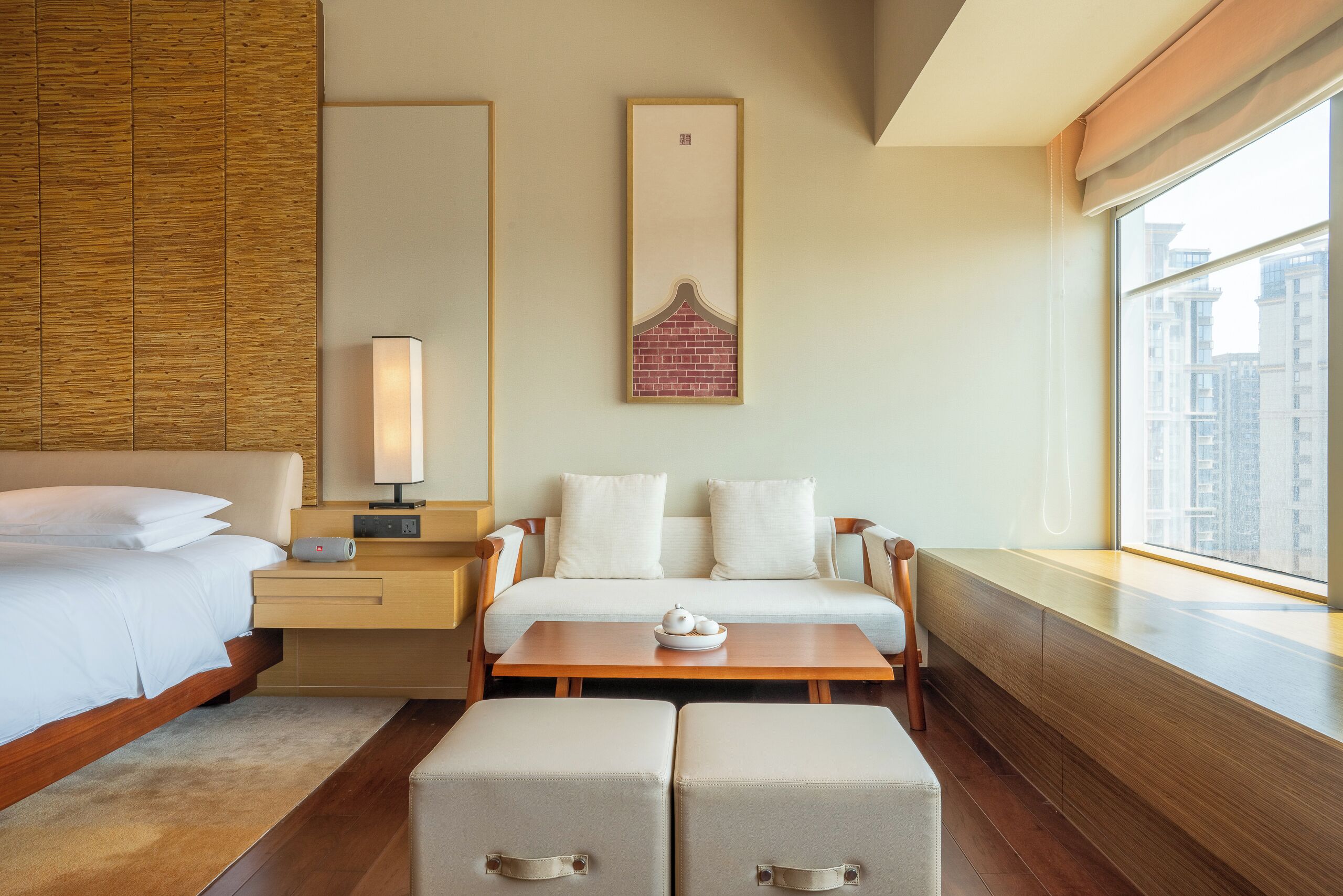 A modern hotel room with a bed, a sofa, a bedside table with a lamp, a small table with a teapot, and a window with a city view.