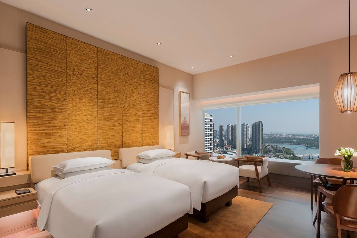 A hotel room with two beds, a desk, a chair, a table, and a large window with a city view.