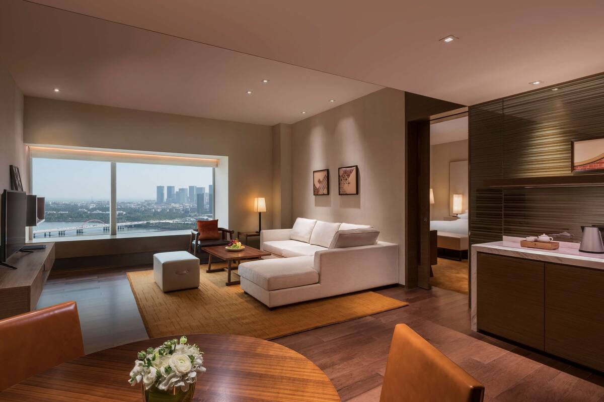 A modern living room with a large window, a white sofa, a dining table with chairs, and a kitchen area.