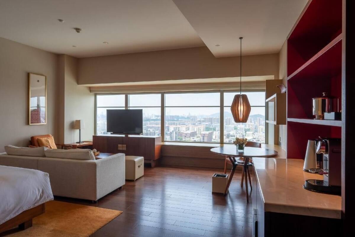A modern hotel room with a large window, a sofa, a bed, a dining table, and a TV.