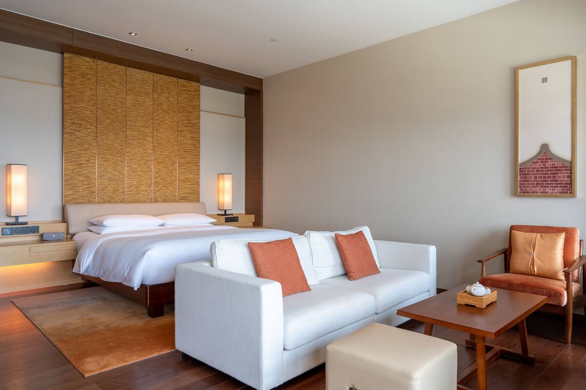 A hotel room with a bed, a sofa, a chair, a small table, and a nightstand with a lamp.